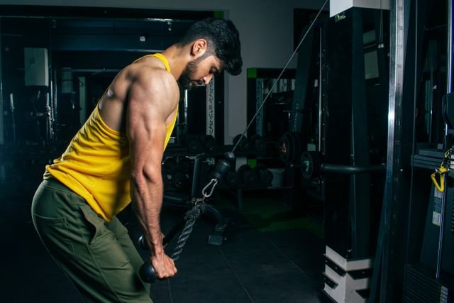 Home A fit man in a yellow tank top exercises using a cable machine in a gym setting.